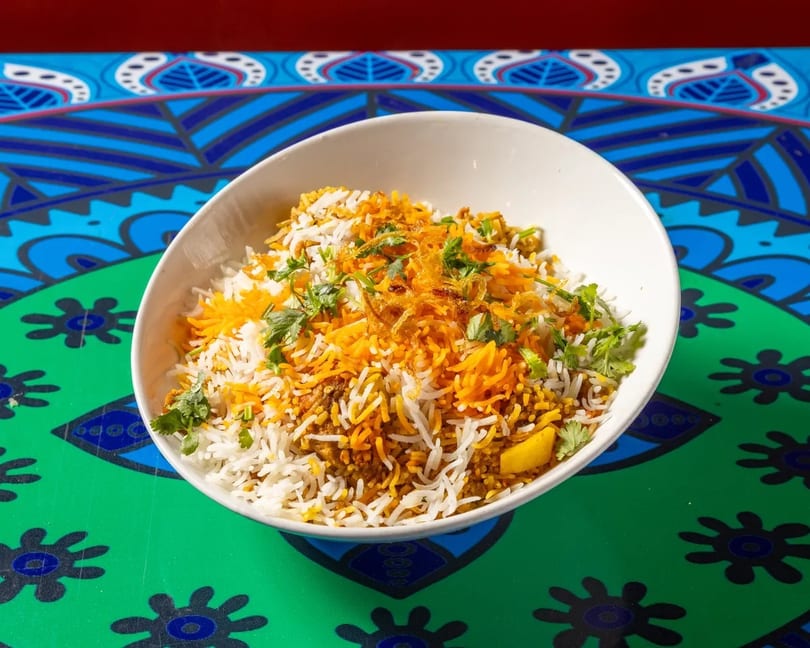 A white bowl of biryani with orange and white rice, cilantro, and fried onions on a colorful table.
