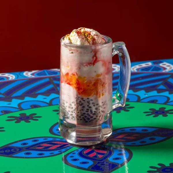 A colorful falooda dessert with ice cream, fruit, and basil seeds in a glass mug on a patterned table.