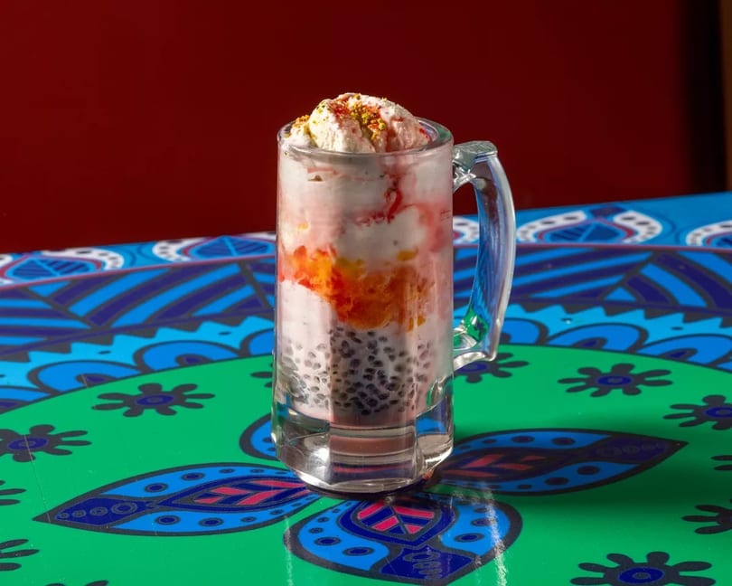 A colorful falooda dessert with ice cream, fruit, and basil seeds in a glass mug on a patterned table.