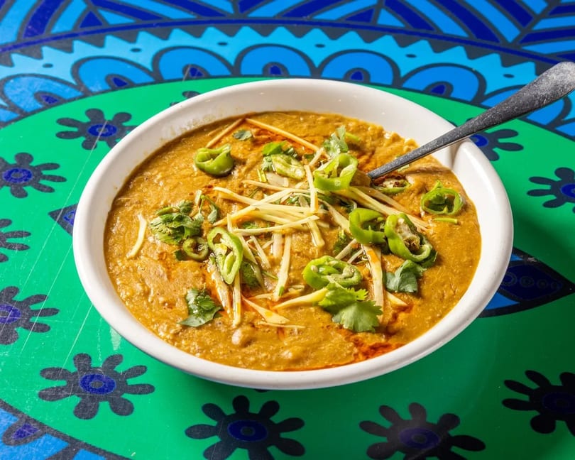 A bowl of haleem garnished with green chilies, ginger, and cilantro on a colorful table.