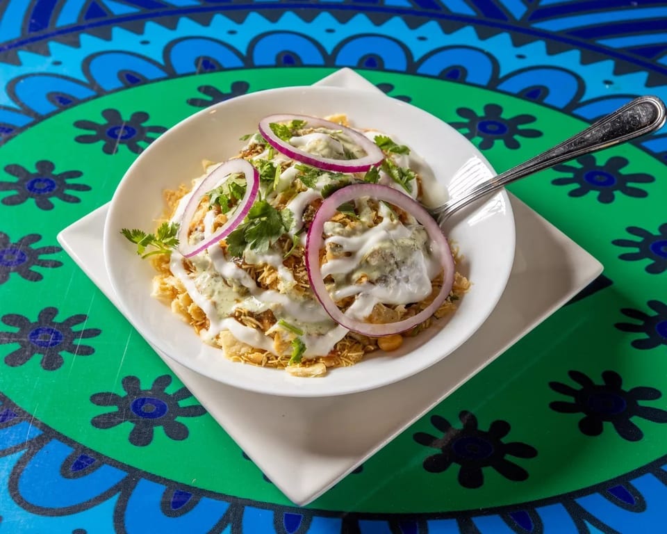 A bowl of Indian chaat with yogurt, red onions, and cilantro on a colorful table.