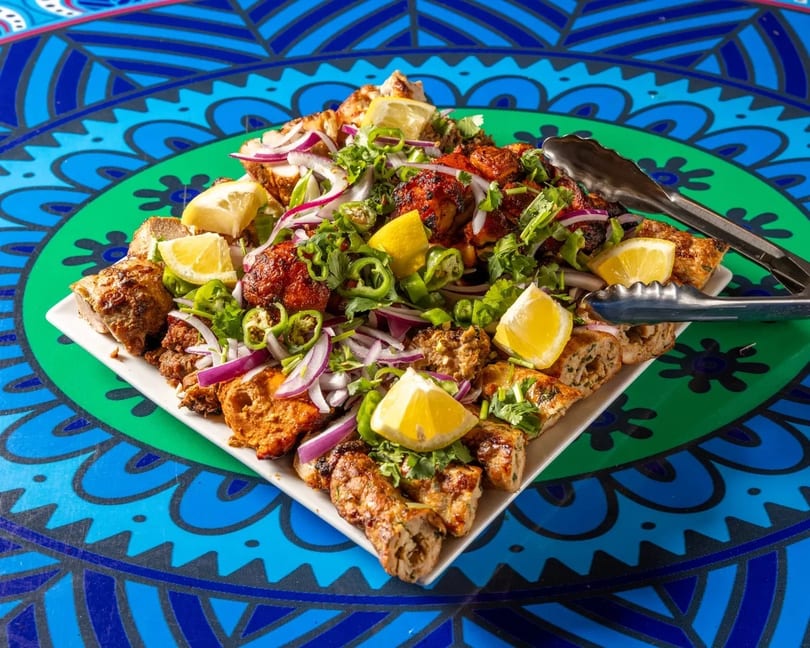 A large platter of grilled meats, onions, peppers, and lemon wedges on a vibrant blue and green table.