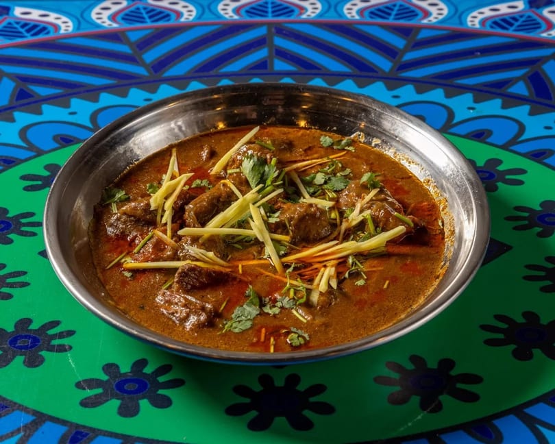Rich brown curry with meat, garnished with fresh ginger and cilantro, in a metal bowl.
