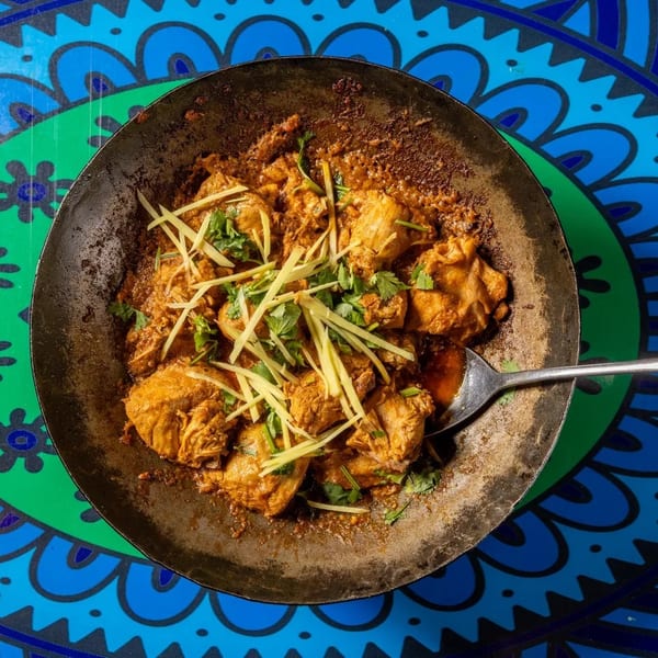 Chicken karahi, garnished with ginger and cilantro, in a metal pan on a blue and green patterned table.