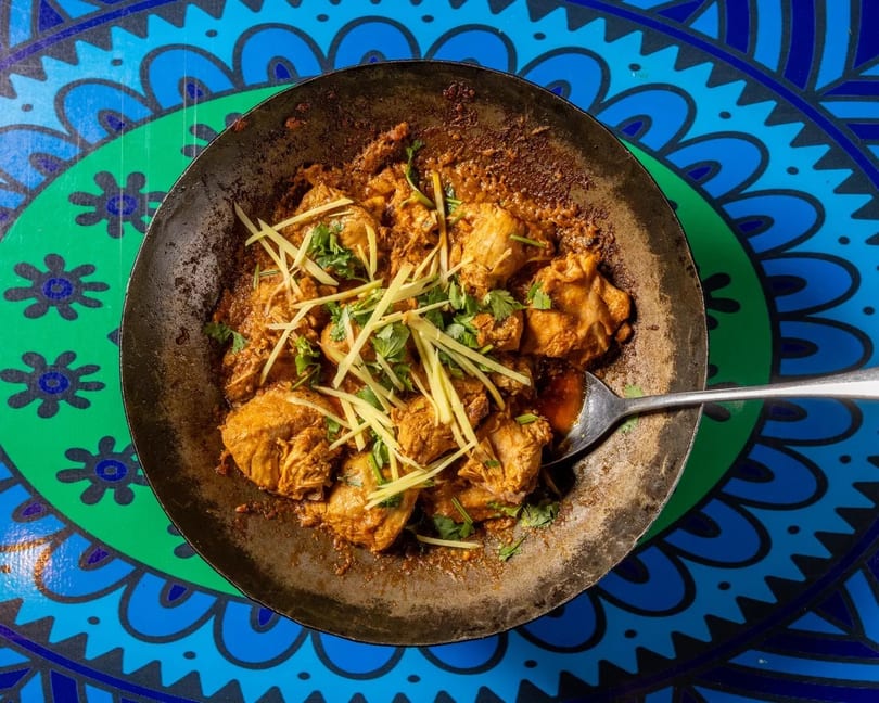 Chicken karahi, garnished with ginger and cilantro, in a metal pan on a blue and green patterned table.