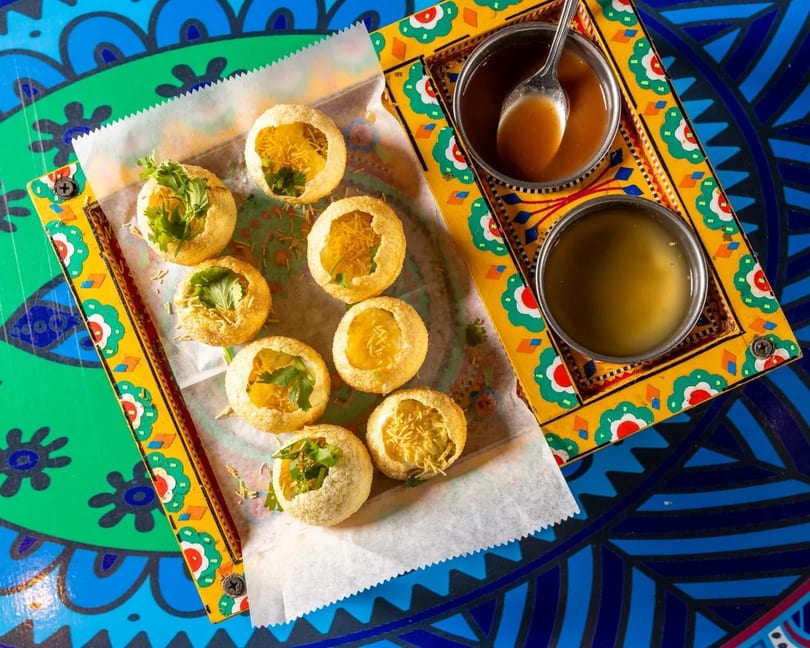 Overhead view of pani puri with two dipping sauces on a colorful patterned table.