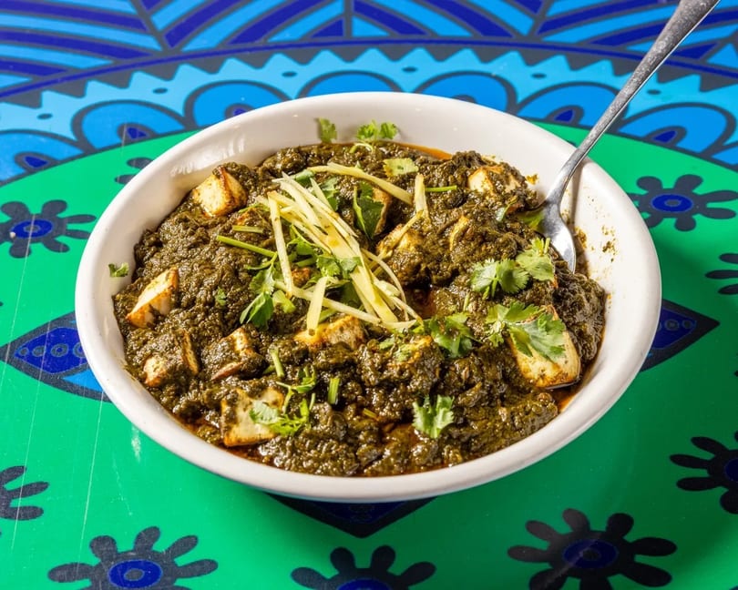 A white bowl of palak paneer with paneer cubes, ginger, and cilantro on a colorful patterned table.