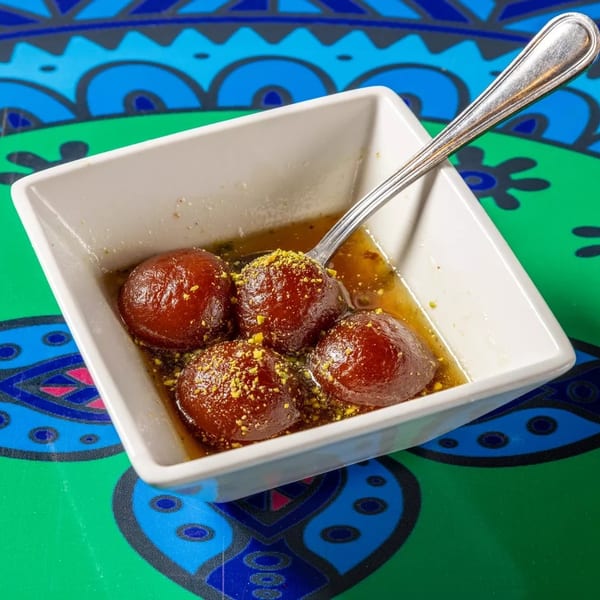 Four gulab jamun in syrup with pistachio sprinkles, served in a white bowl on a colorful patterned table.