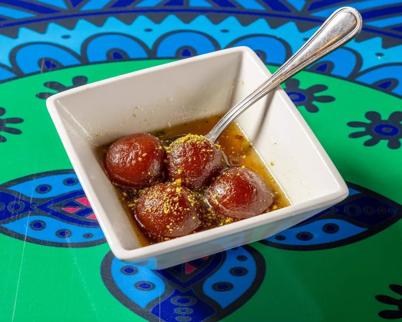 Four gulab jamun in syrup with pistachio sprinkles, served in a white bowl on a colorful patterned table.
