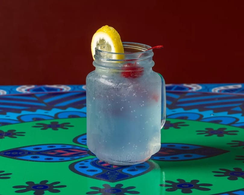 A frosty mason jar drink with a lemon slice and cherry on a colorful patterned table.