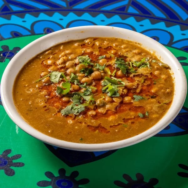A white bowl of chickpea curry with cilantro and chili oil on a vibrant green and blue patterned table.