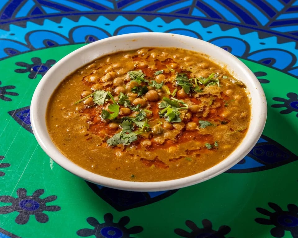 A white bowl of chickpea curry with cilantro and chili oil on a vibrant green and blue patterned table.