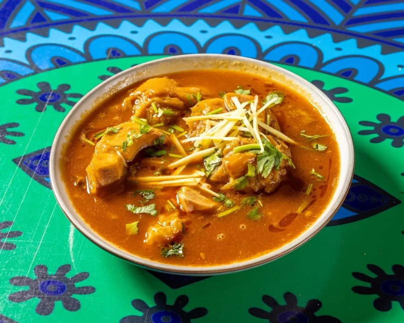 A bowl of rich, orange-brown curry with meat pieces, garnished with cilantro and ginger, on a colorful table.