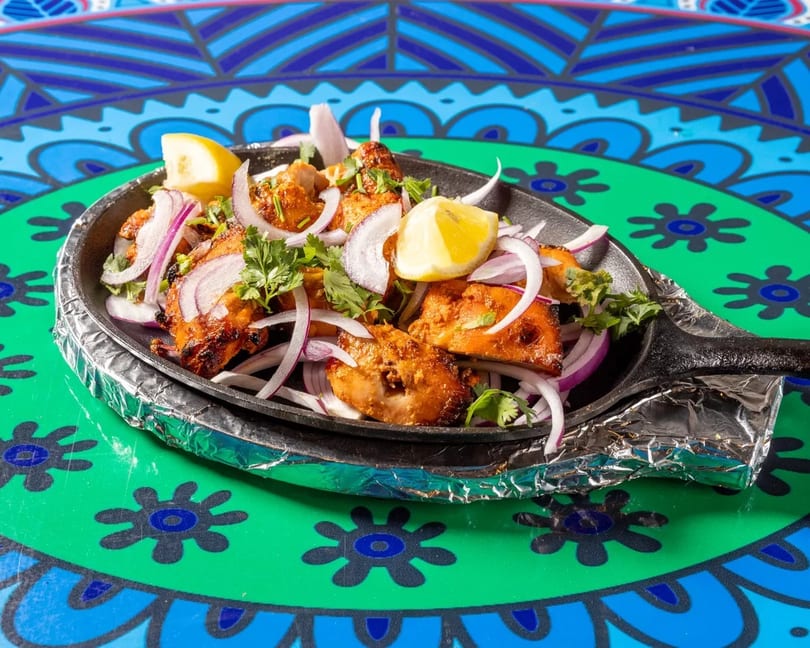 Sizzling chicken tikka with red onions, cilantro, and lemon wedges on a colorful patterned table.